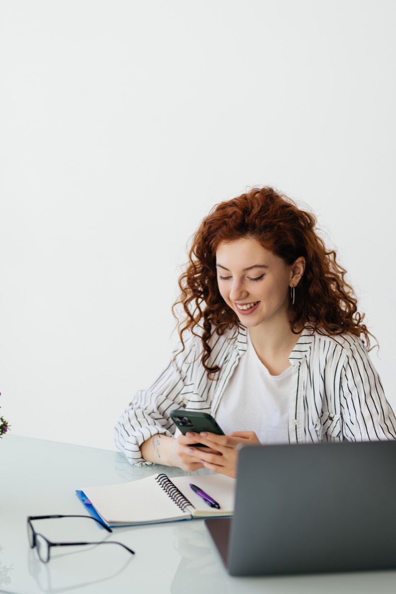 Mulher sorrindo enquanto usa o celular, com caderno e notebook na mesa